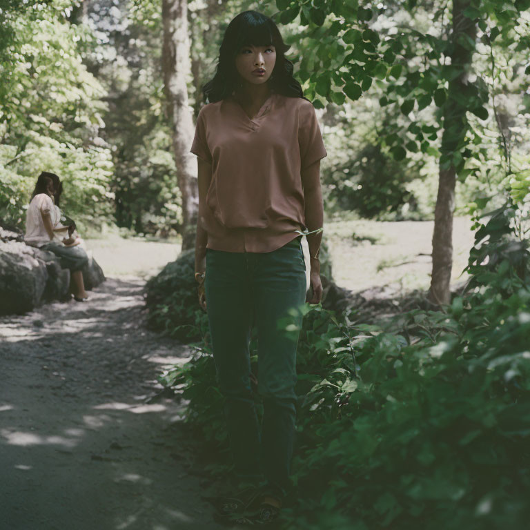 Woman on forest path gazes at camera, another person in background among lush greenery