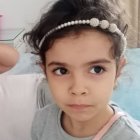 Portrait of young child with brown eyes and pearl headband, arm on cushion, pink candle holder.