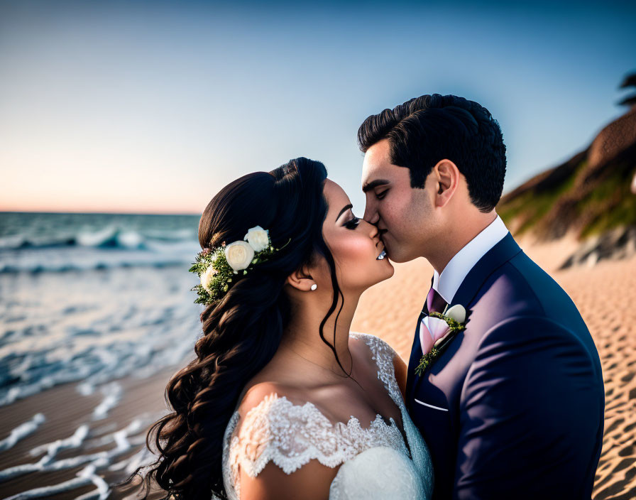 Bride and groom kissing on beach at sunset with ocean backdrop