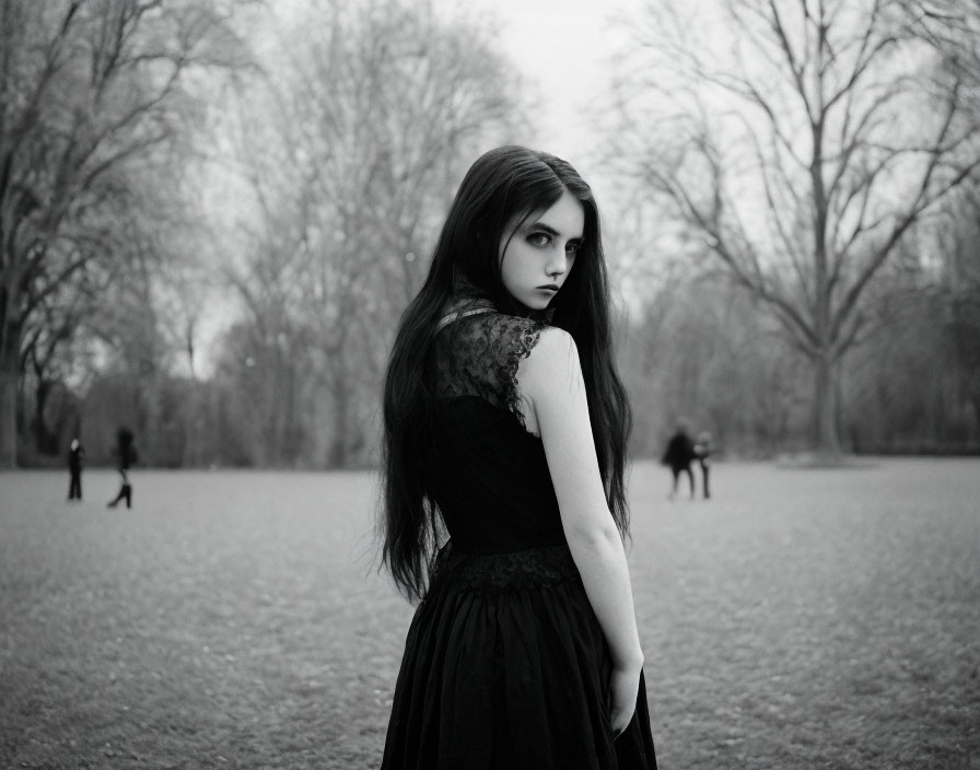 Dark-haired woman in black dress stands in park with pensive expression