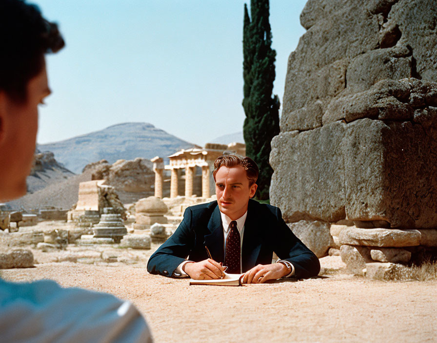 Man in suit writing at ancient ruins with person's head in foreground