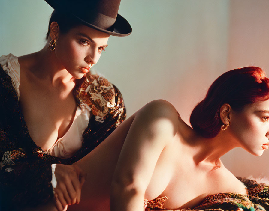 Two women in dramatic lighting: one with black hat and corset, the other reclining thoughtfully