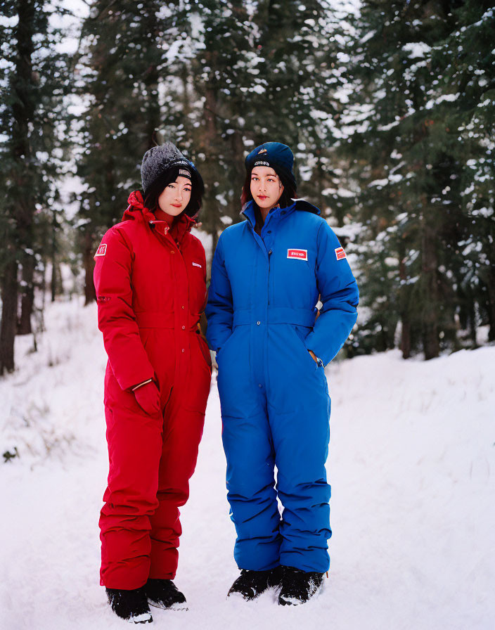 Two people in red and blue winter jumpsuits in snowy forest.