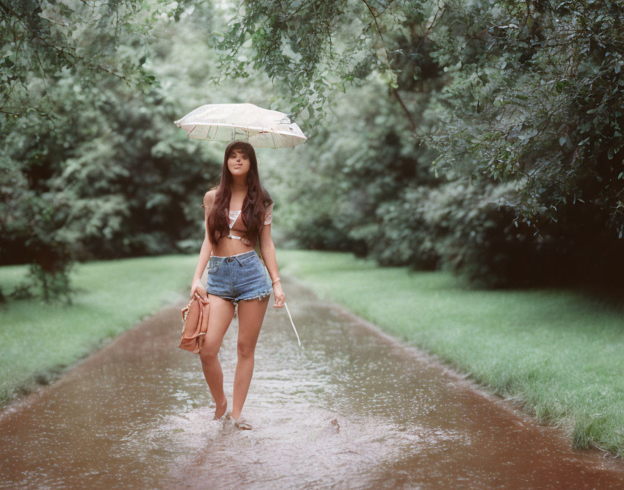 Woman with umbrella walking on wet park path among lush green trees wearing hat and denim shorts.