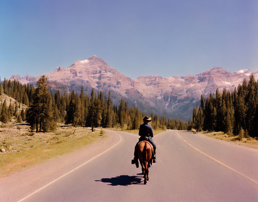 Cowboy riding horse on deserted highway with mountains and blue sky