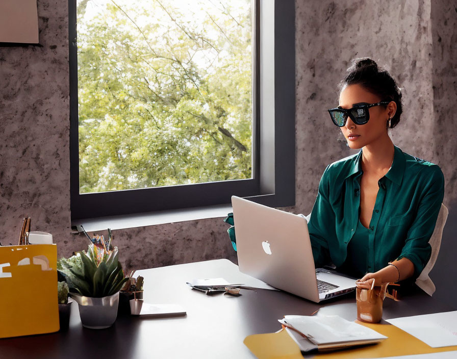 Woman in green blouse works on laptop at desk by window