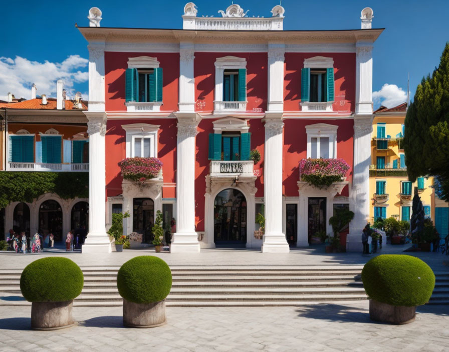 Vibrant red building with white trim, balconies with flowers, and spherical bushes under blue sky