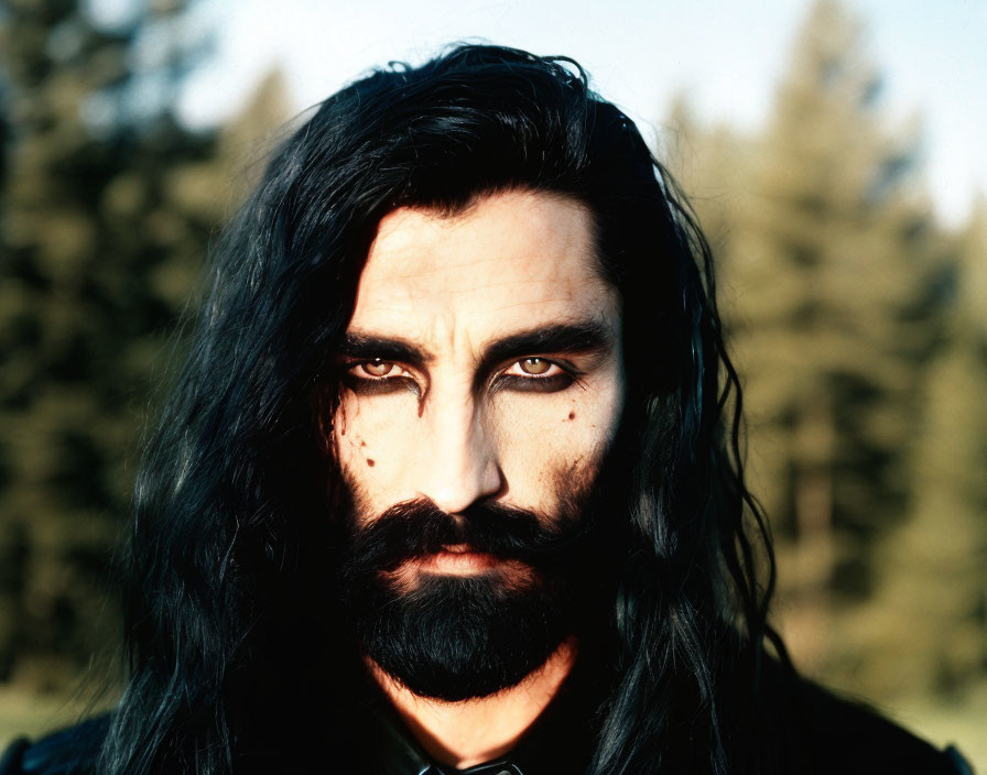 Bearded person with long black hair outdoors among trees