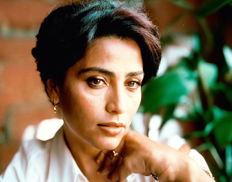 Dark-haired woman with earrings gazes at camera against brick wall.