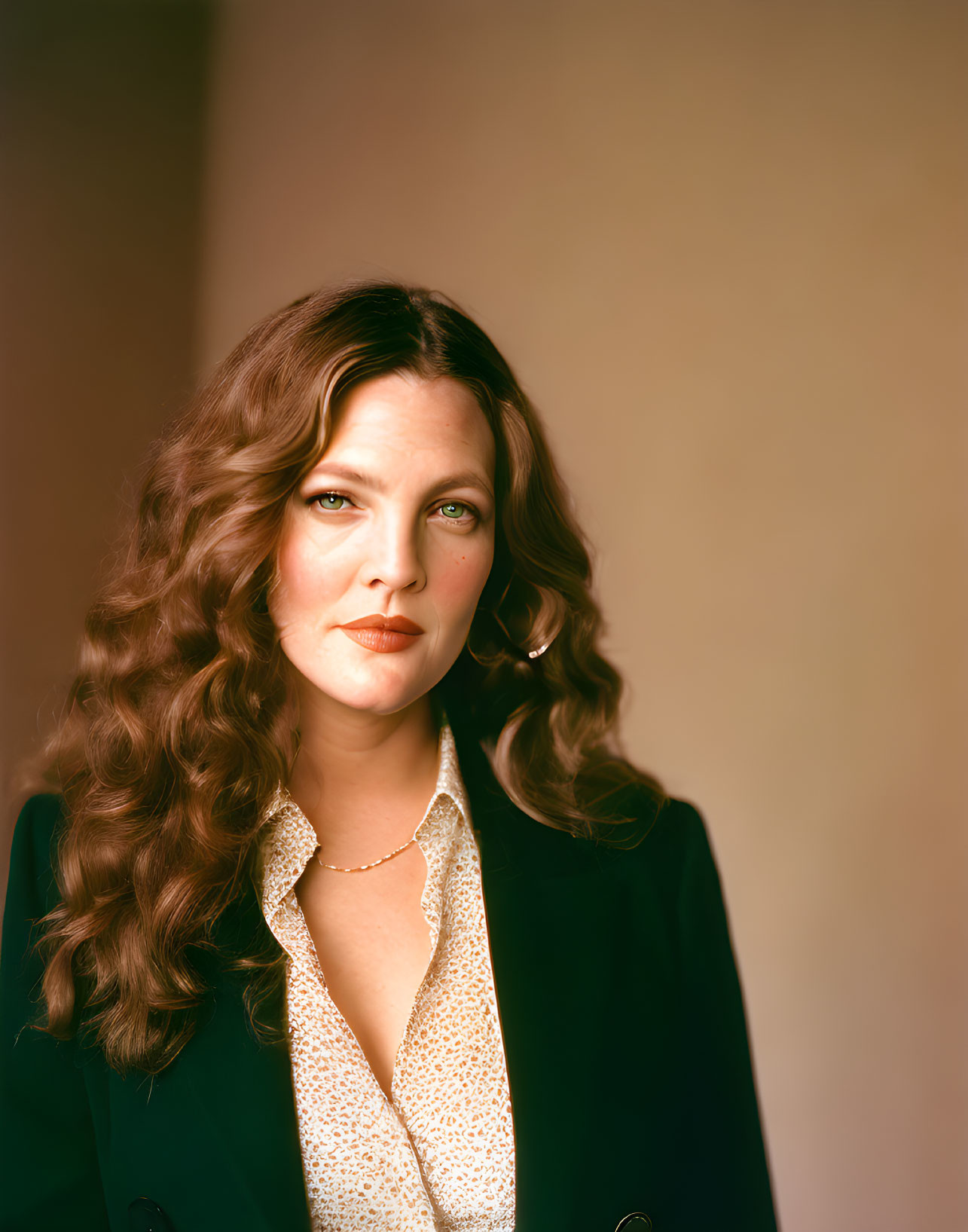 Long-haired woman in dark blazer and patterned blouse gazes at camera.