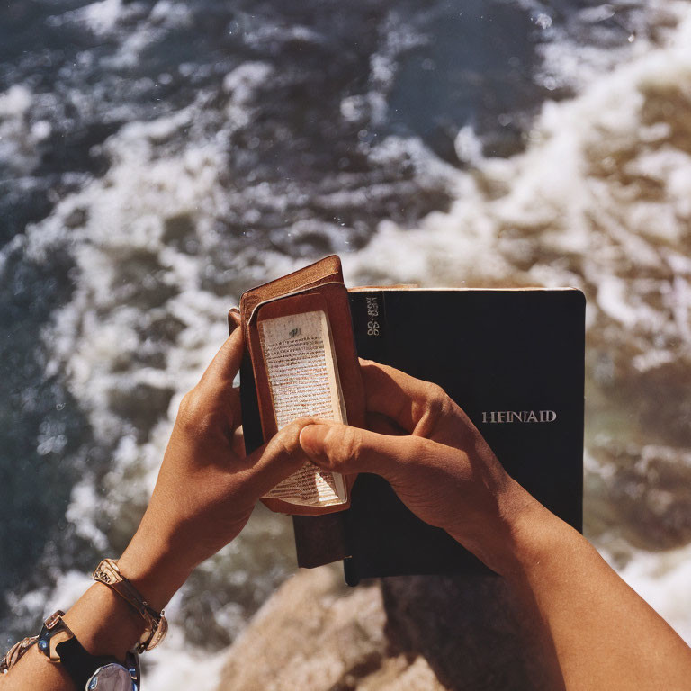 Person reading small book near water with sunlight and shadows on pages.