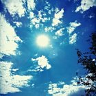 Scattered clouds in vibrant blue sky with bright sun and tree branches