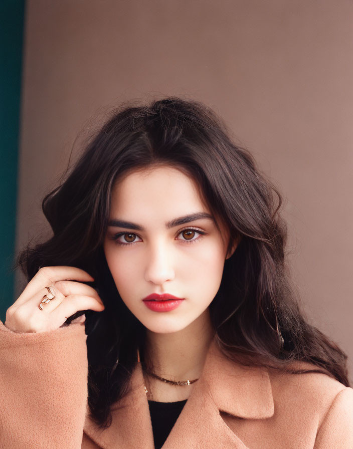 Portrait of woman with dark hair, bold eyebrows, red lipstick, beige coat, delicate necklace on blue