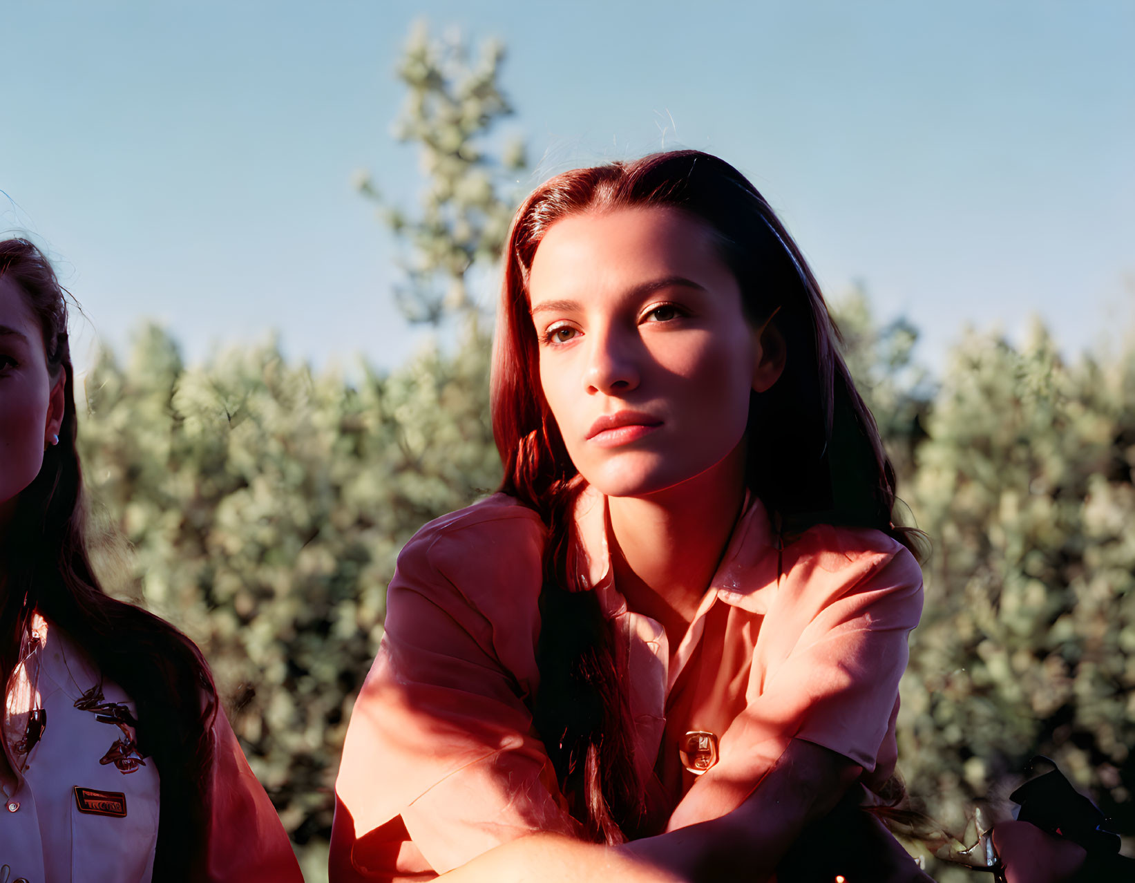 Young woman with long hair in sunlight with another person and greenery in background.