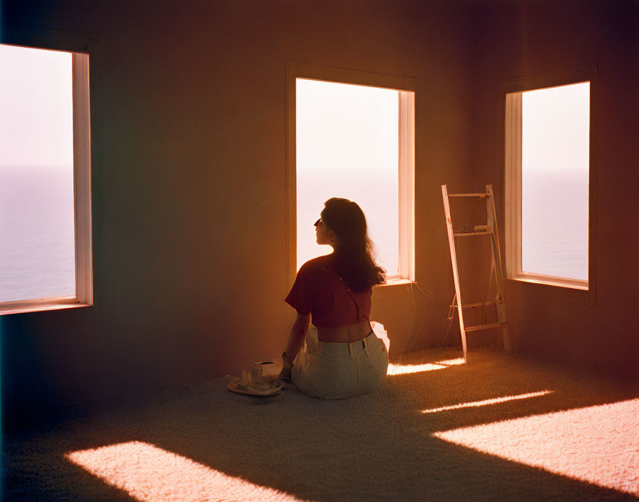 Person sitting on carpeted floor looking out window at calm sea during sunset with warm light in room.