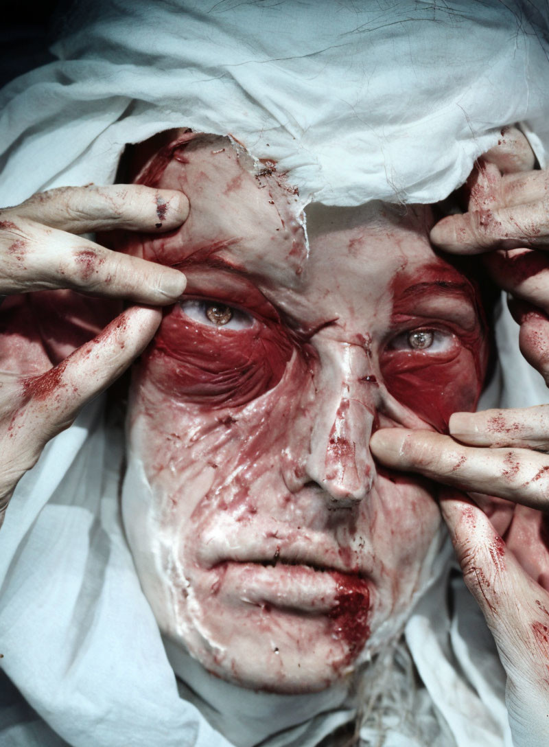 Blood-stained person gazes through white fabric, intense and haunting.