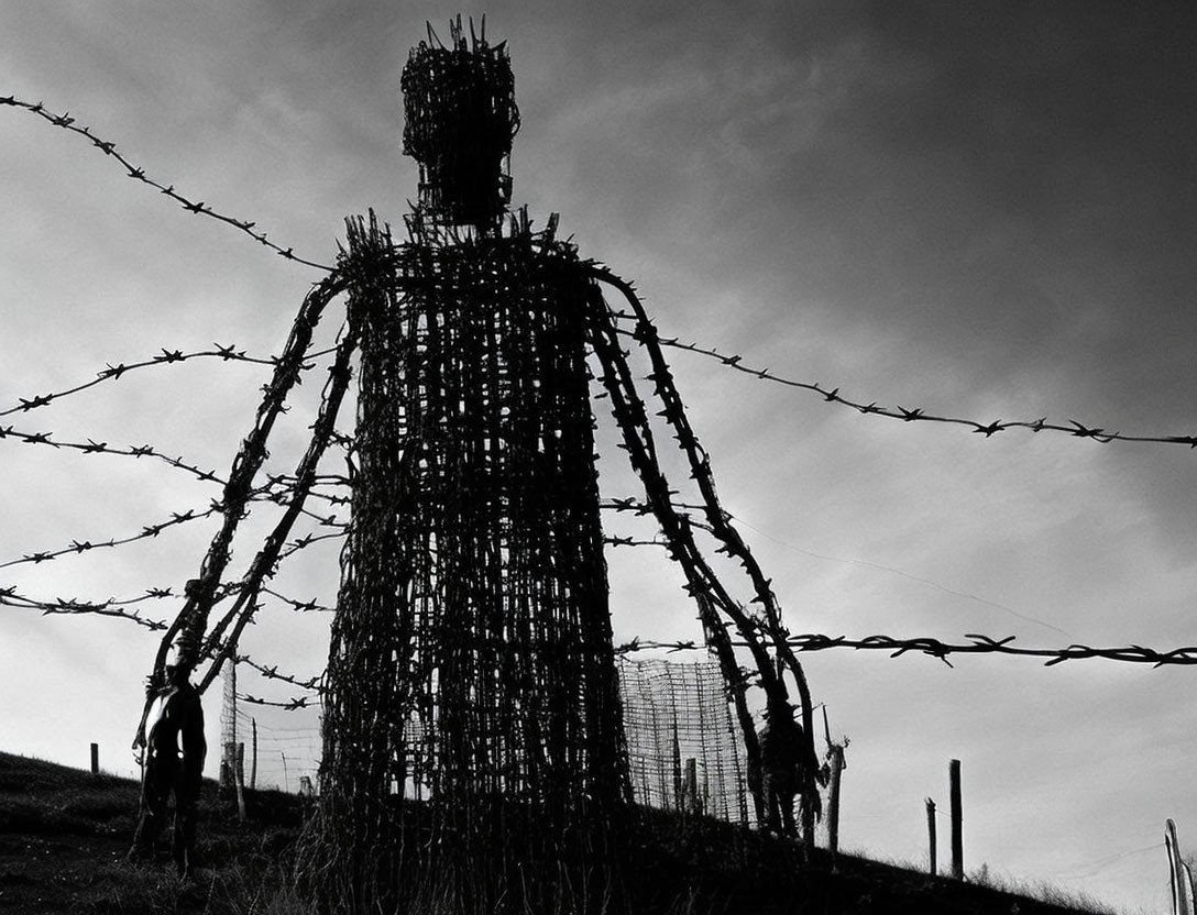 Wire silhouette of large figure behind barbed wire under gloomy sky