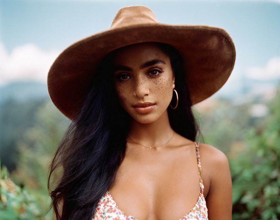 Freckled woman in brown hat and floral outfit against mountainous backdrop