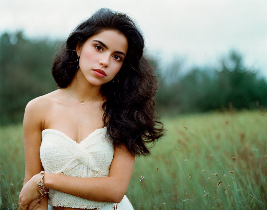 Dark-haired woman in white dress standing in field with crossed arms