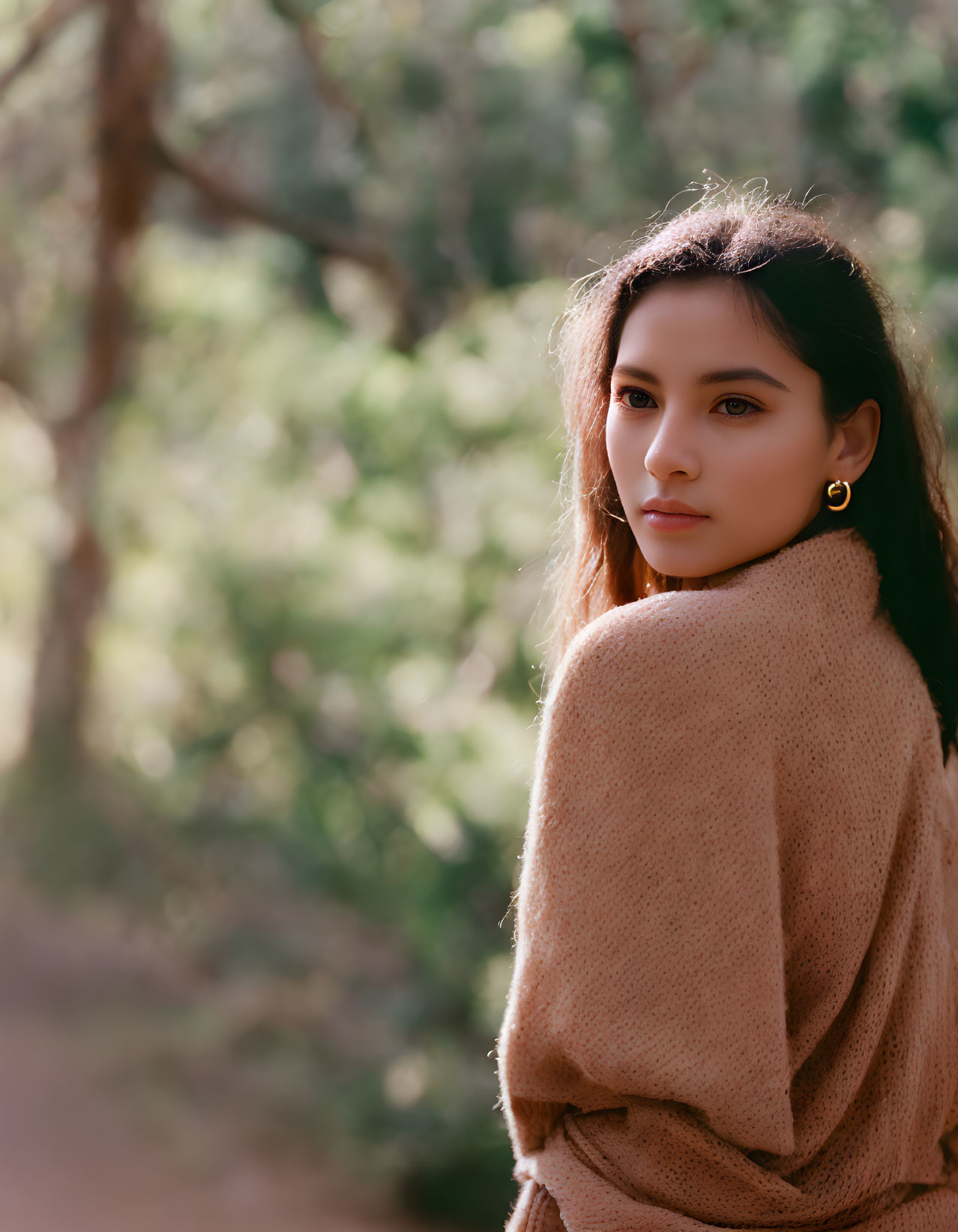 Long-haired person in brown sweater glances over shoulder in nature setting