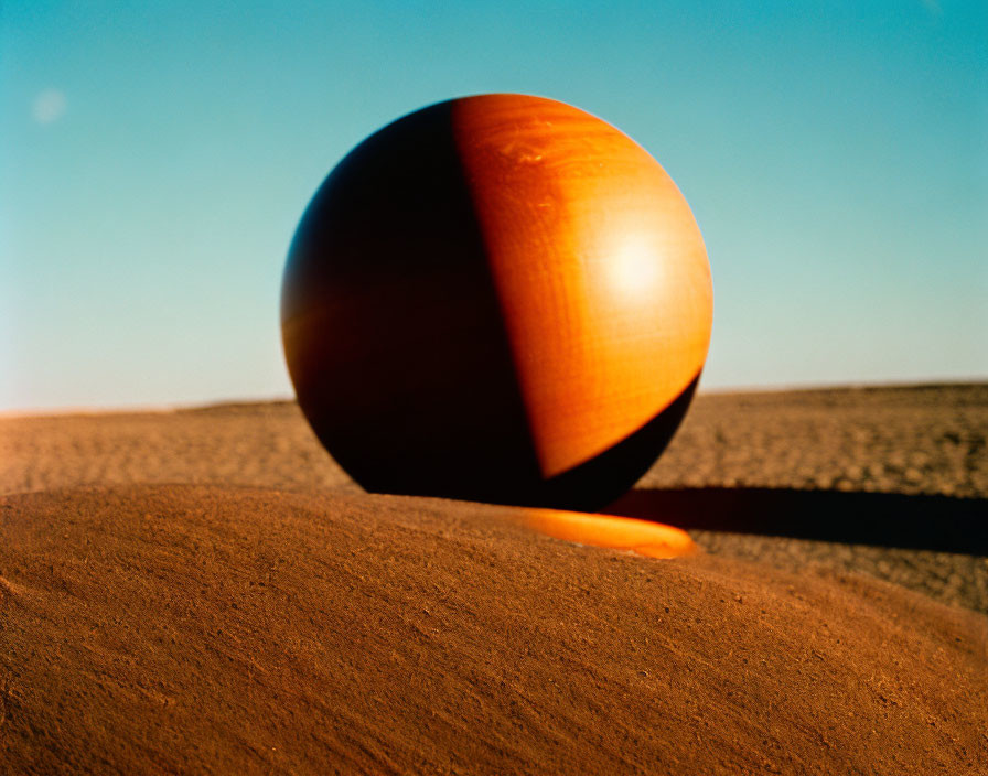 Orange Sphere on Sandy Surface with Long Shadow Against Blue Sky