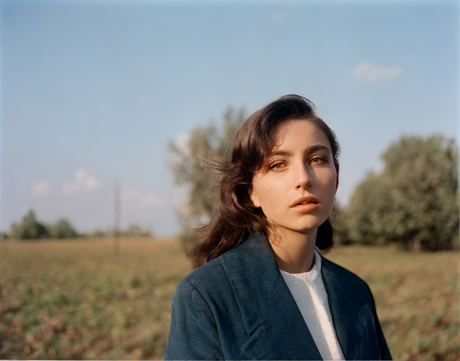 Woman in windswept hair, blue blazer, white shirt, gazing in open field.