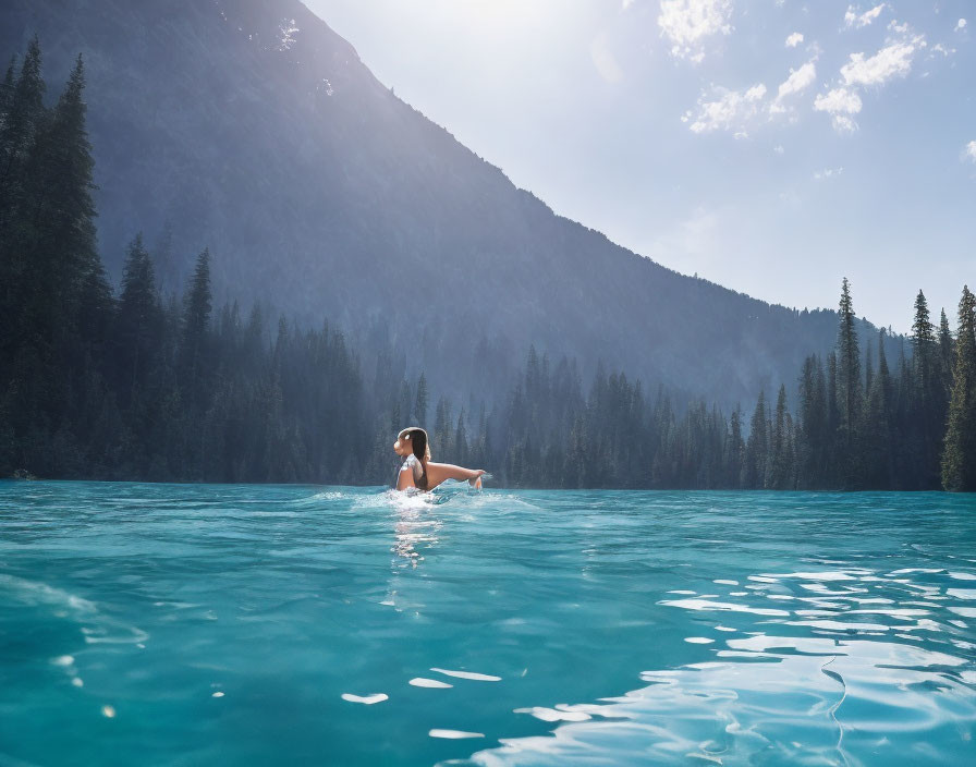 Person swimming in clear blue mountain lake with forested slopes and sunny skies