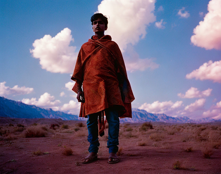 Person in patterned shawl in desert landscape with mountains and cloudy sky