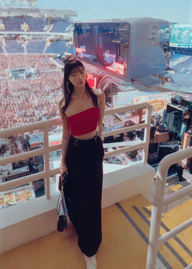 Woman in red top and black pants in stadium with large screen and crowd.