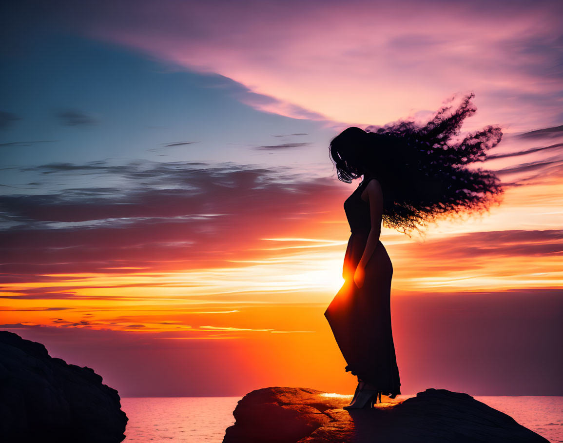 Woman's silhouette with flowing hair on rock at sunset.