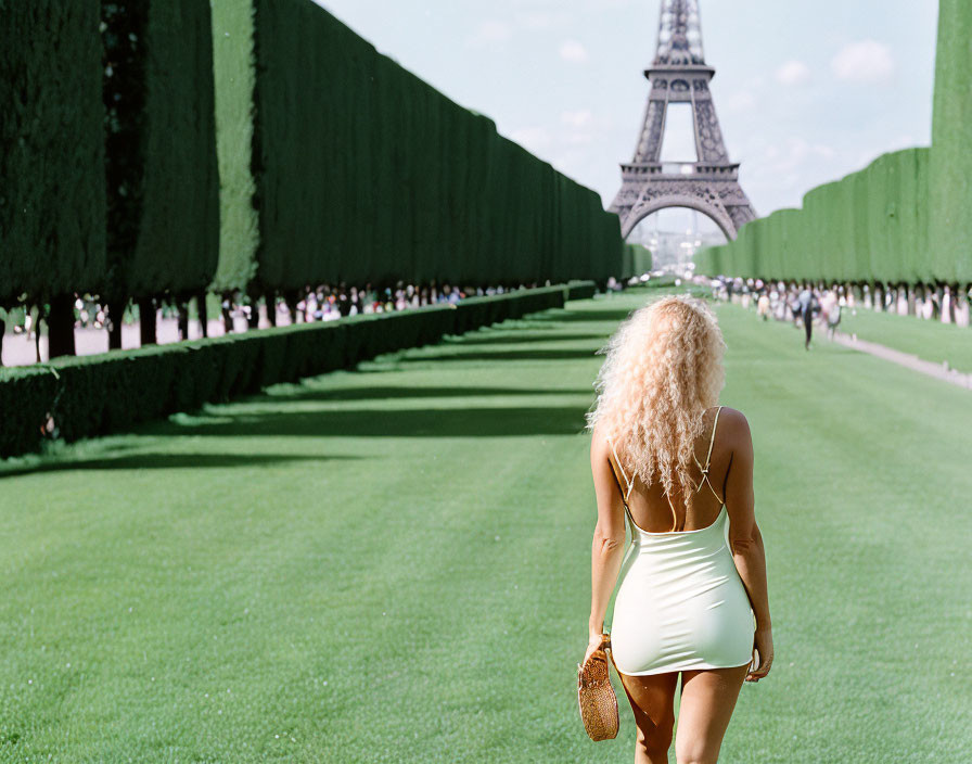 Blonde woman in light dress walking to Eiffel Tower on green lawn