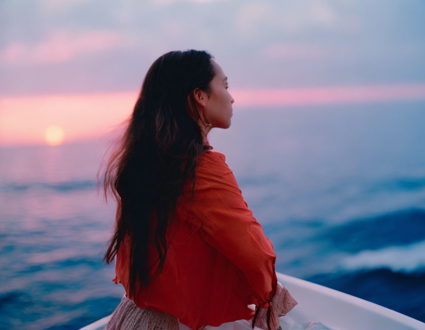 Woman in orange top gazes at sunset over ocean from boat deck