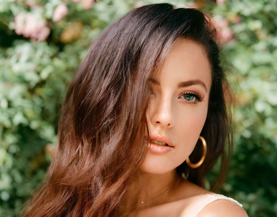 Brown-haired woman with green eyes in hoop earrings surrounded by green foliage and pink flowers