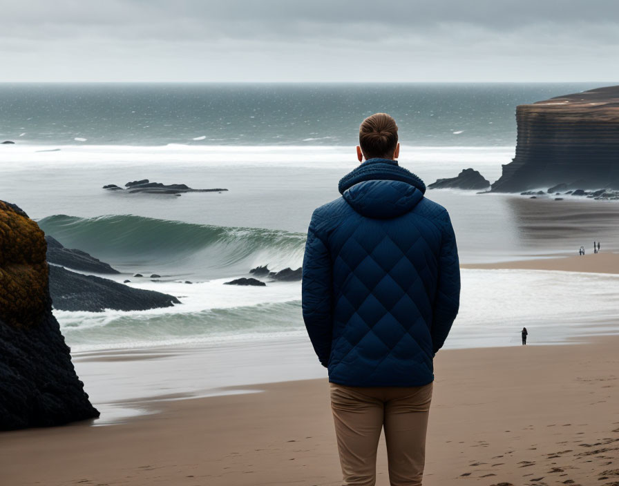 Person in Blue Jacket on Beach Overlooking Waves and Cliffs