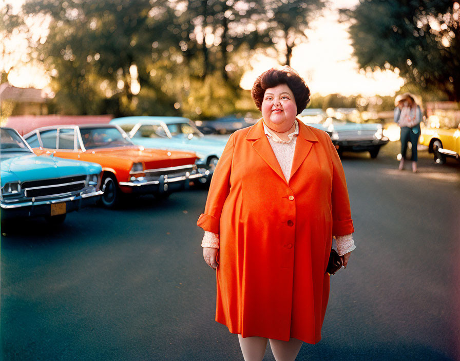 Smiling woman in bright orange coat on sunny street with vintage cars.