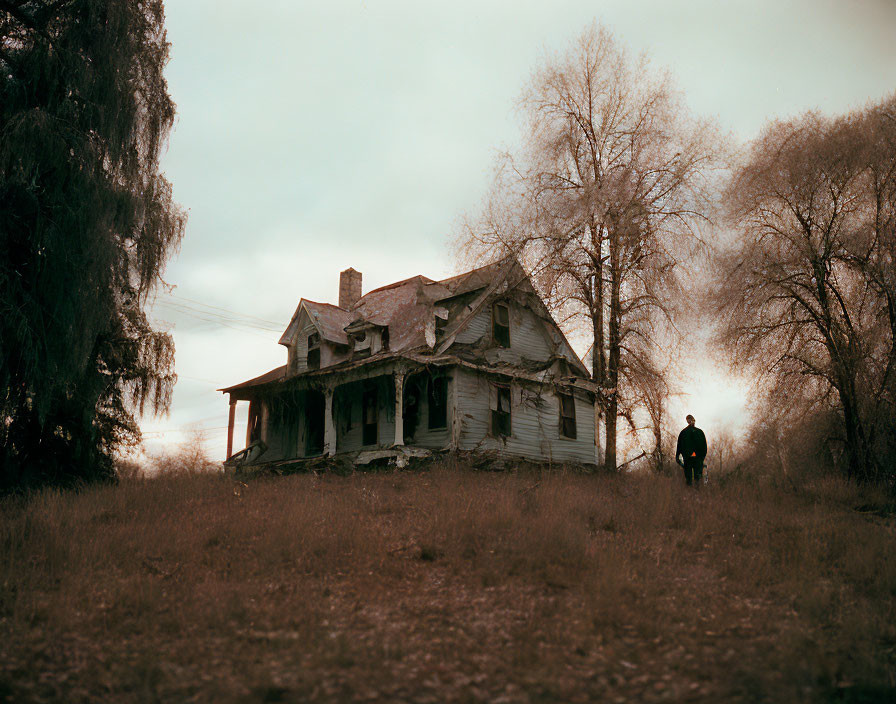 Abandoned two-story house with porch, person, bare trees, and cloudy sky