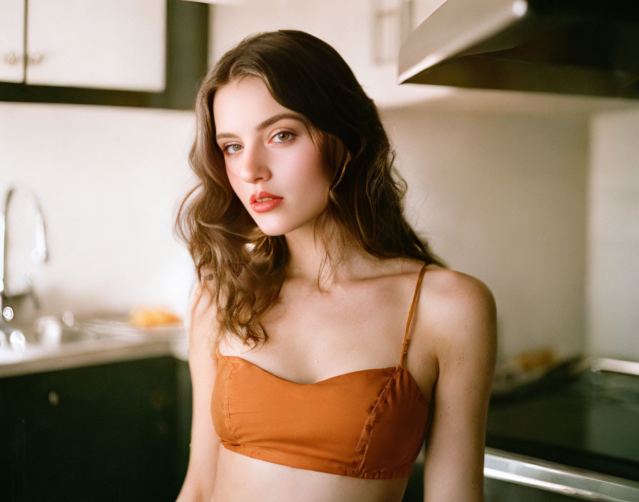 Young woman with wavy brown hair in rust camisole poses in kitchen setting