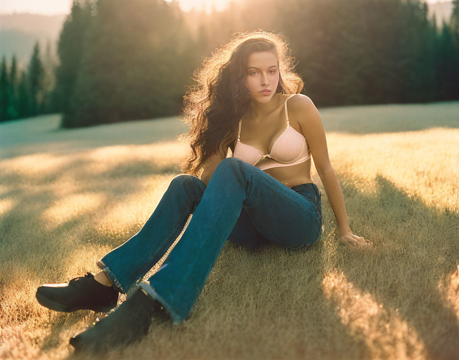 Woman sitting on grass at sunset in jeans, light top, boots, wavy hair.