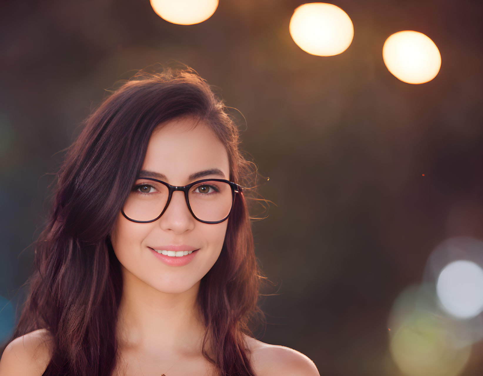 Smiling woman with glasses and long brown hair in blurred background.