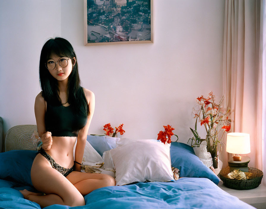 Woman with glasses sitting on blue bed near nightstand with lamp, flowers, and cityscape painting.