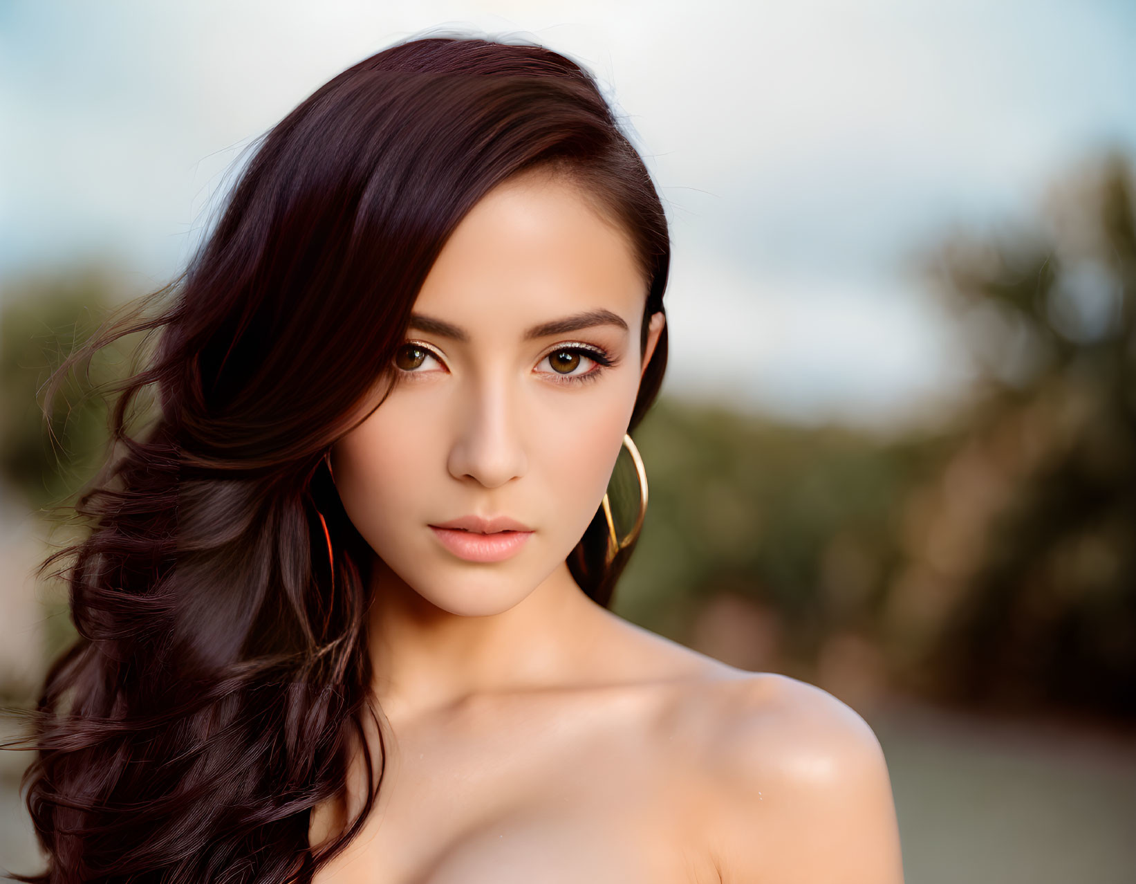 Portrait of woman with dark wavy hair, gold earrings, subtle makeup, and bare shoulder against blurred