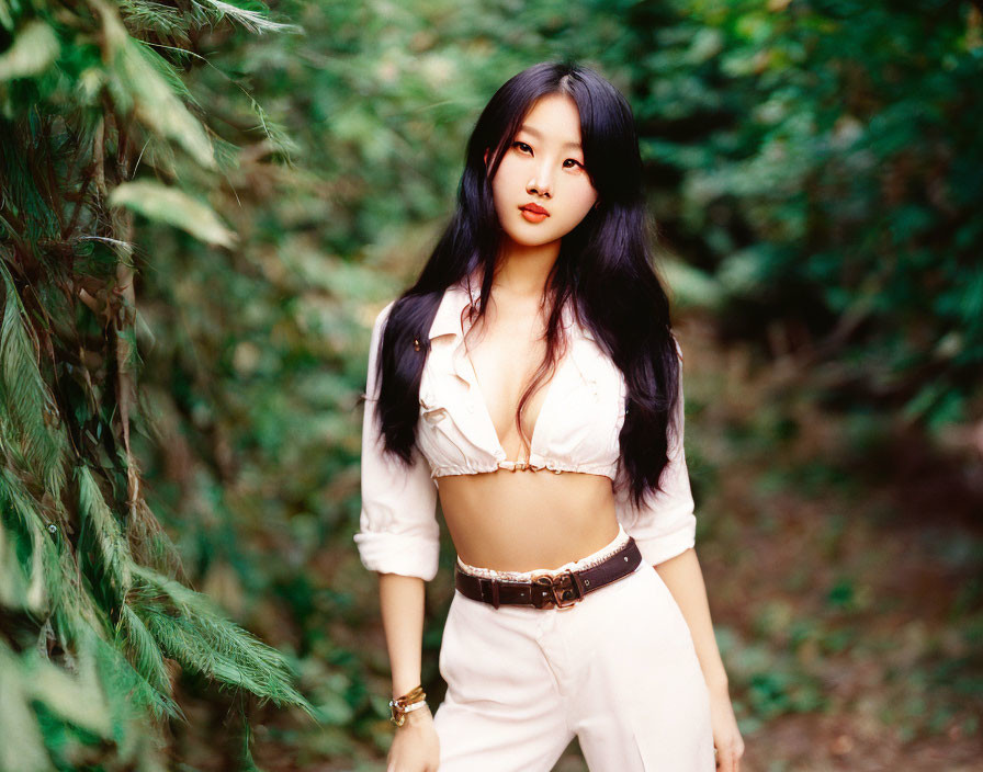 Woman with long black hair in white cropped shirt and high-waisted pants in lush green foliage