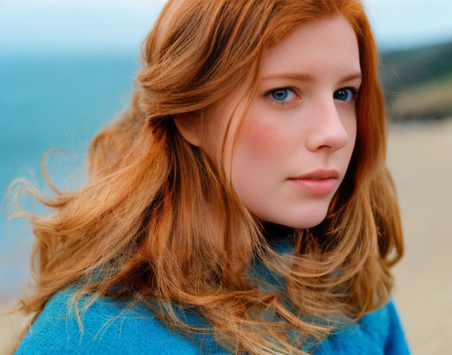 Young woman with auburn hair in blue sweater smiles against beach backdrop