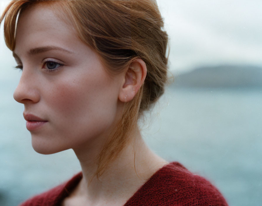 Pensive young woman with auburn hair in red sweater against seascape.