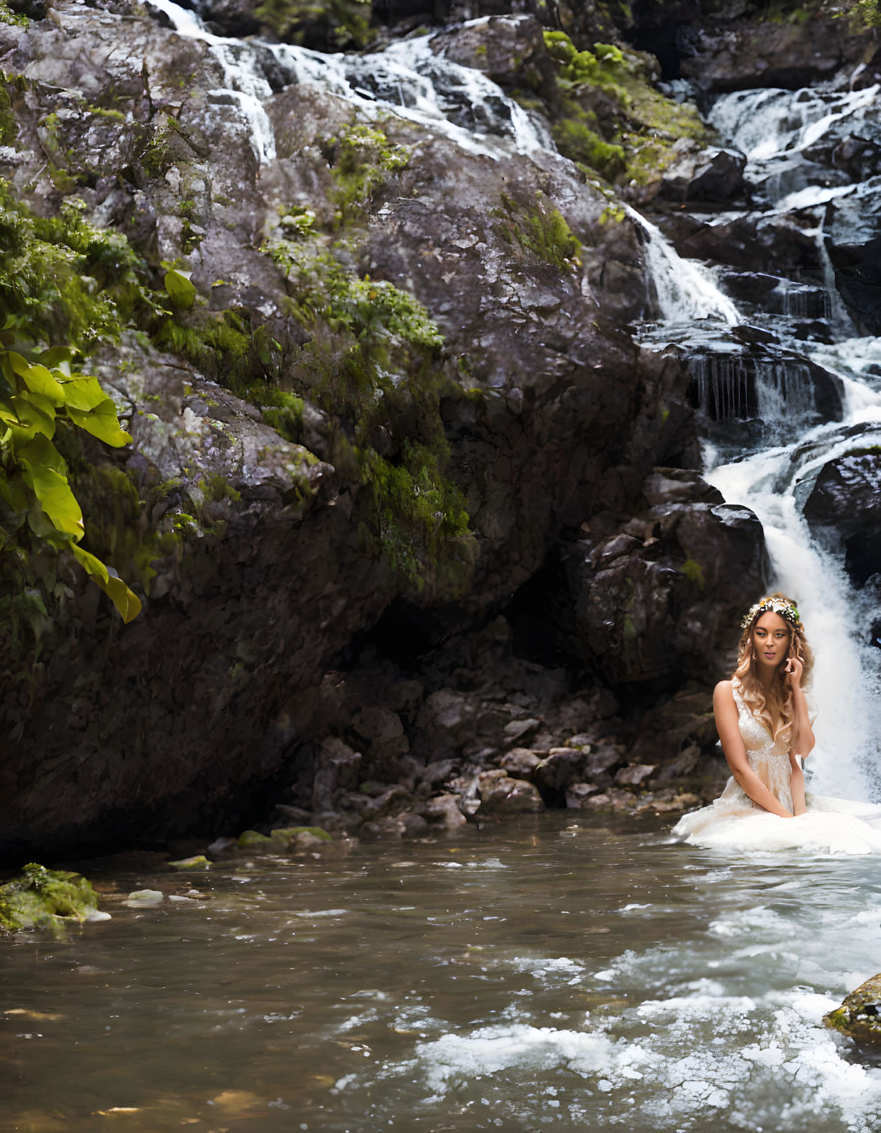 Person in white dress with floral headpiece in forest stream with waterfall.