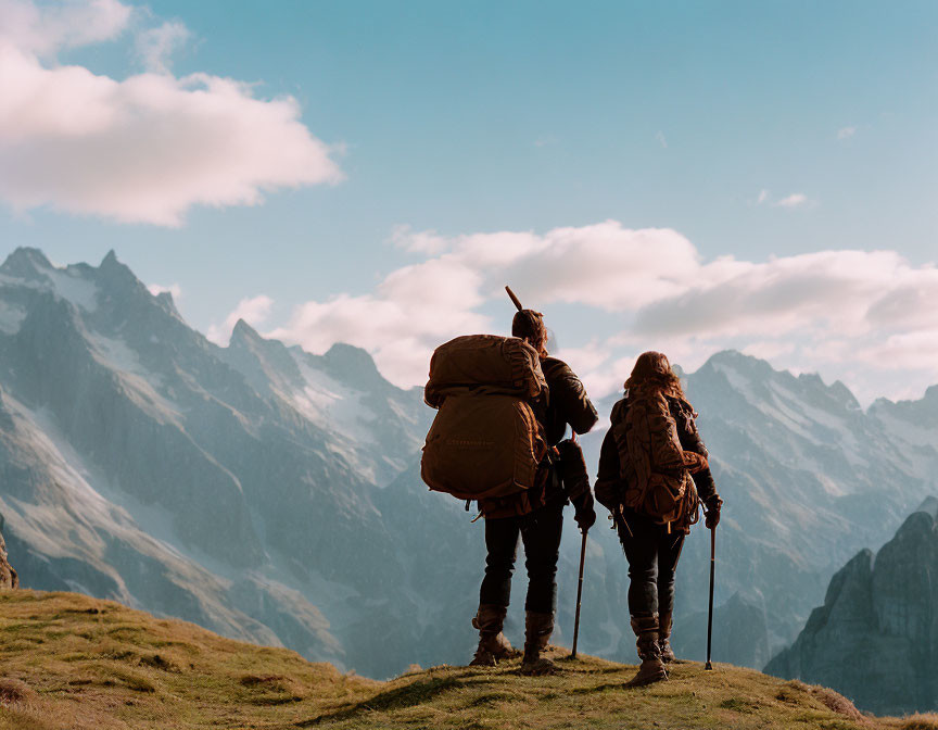 Hikers with backpacks on mountain ridge, sharp peaks under soft cloudy sky