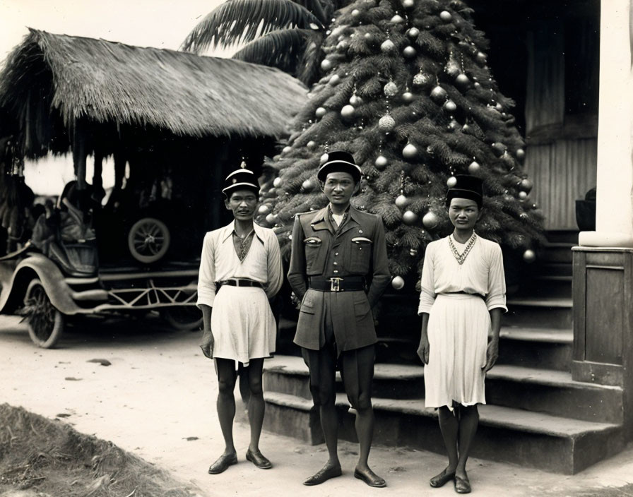 Three people pose by Christmas tree near tropical buildings and vintage car