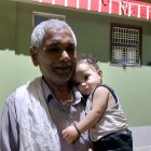 Elderly man with white mustache hugging young child in front of colorful house