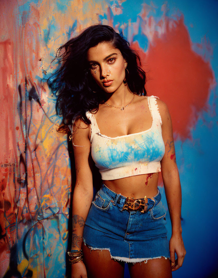 Dark-haired person with tattoos in white crop top and denim shorts on vibrant paint-splattered backdrop