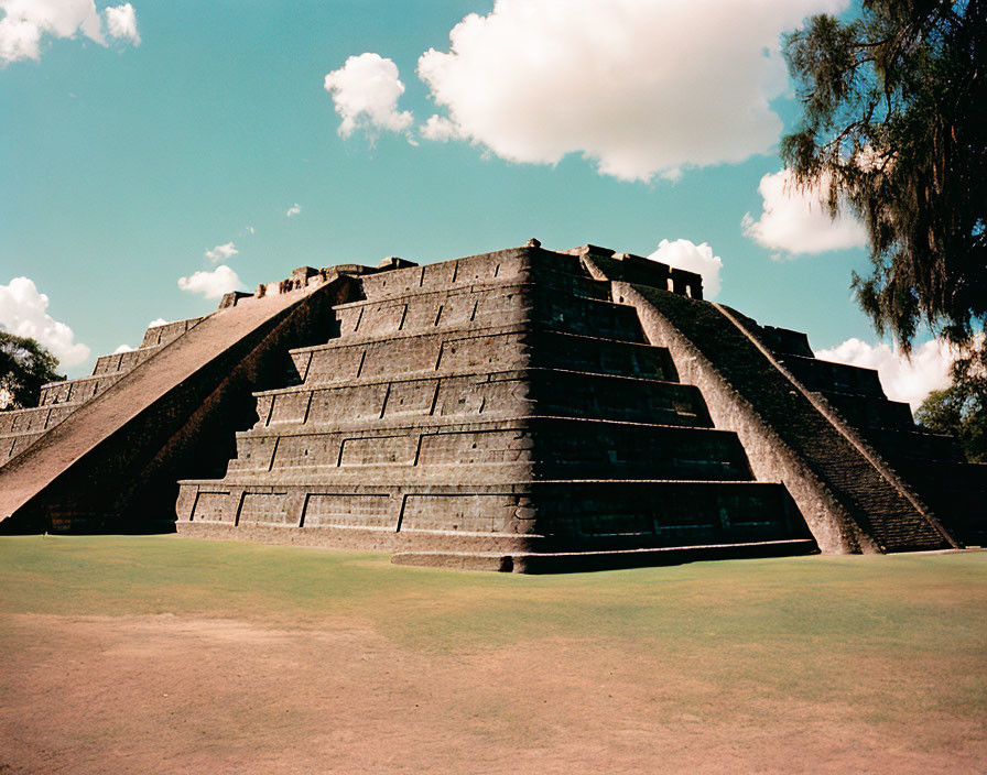 Pyramid with staircases under blue sky, green grass, and tree.